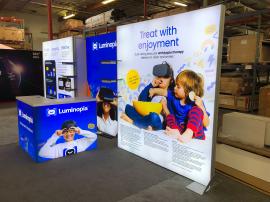 RENTAL: RE-2083 10’ x 20’ Inline Design with (3) Single-Sided Lightboxes, (6) Black Laminated Product Shelves, RE-1572 L-Shaped Backlit Reception Counter with White Laminate Top, and SEG Fabric Graphics.