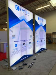 RENTAL:  Island Design with (2) 8' Wide x 12' High Double-Sided Lightboxes, (2) RE-1575 White Laminated Workstation Counters, (6) Large Monitor Mounts, (2) L-Shaped Hanging Signs, RE-1572 L-Shaped Backlit Reception Counter, SEG Fabric Graphics, and Pillow