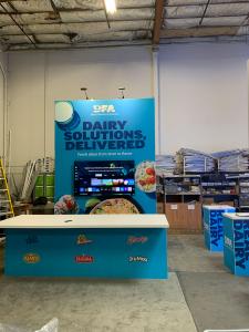 RENTAL: Modified RE-9084 Island Design with Gravitee Storage Closet with Locking Door, (3) 3 ft. Wide x 15 ft. High Double-Sided Lightboxes, (3) RE-1219 Square Workstation Counters, Large Rectangular Counter, (6) LED Arm Lights, 10 x 10 x 3 Square Hanging